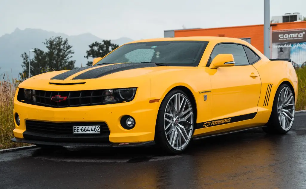 A yellow chevrolet coupe with two black stripes on rained asphalt.