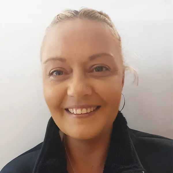 Close-up of a smiling woman with blonde hair tied back, wearing hoop earrings and a black jacket.
